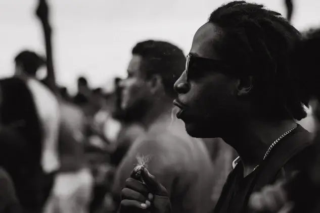 Sepia-style photo of a black man smoking a joint wearing black sunglasses in a crowd of people at a music festival on 4/20